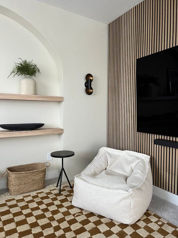 A warm, contemporary living room corner featuring a sculptural white lounge chair, small black side table, woven basket, checkerboard rug, arched built-in shelving, and a vertical timber slat feature wall surrounding a wall-mounted television. The palette is soft white, light timber, black, and brown with refined, tactile finishes.