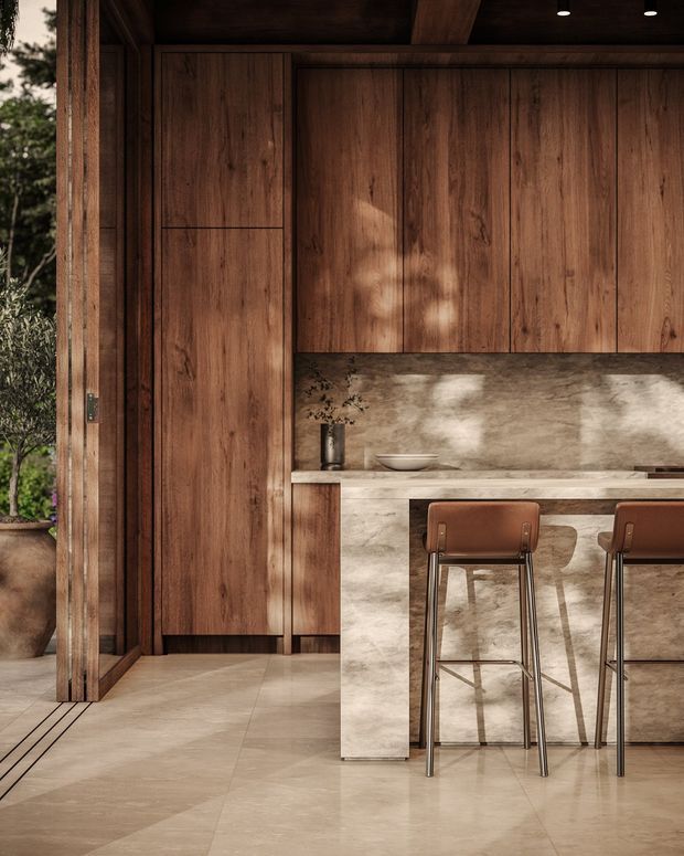 Warm, refined kitchen interior with floor-to-ceiling dark timber cabinetry, a large stone island and matching stone splashback, integrated low-profile lighting, and leather bar stools. A timber-framed opening connects the kitchen to planted exterior space, while natural light creates dappled shadows across the joinery and stone surfaces.
