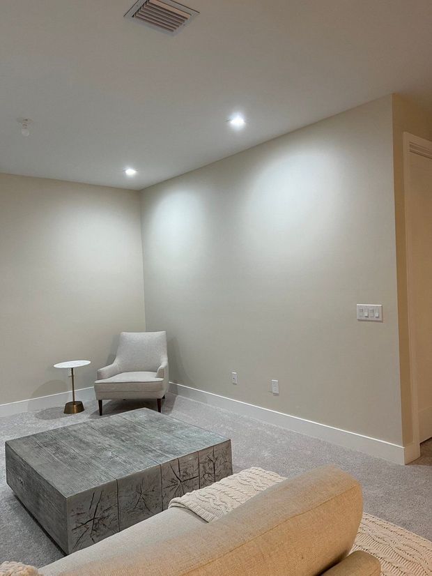 A quiet, neutral-toned living room with beige painted walls, recessed ceiling lights, carpeted flooring, and a minimalist seating arrangement. A pale upholstered sofa faces a large distressed grey wood coffee table, with a light armchair and small round side table positioned in the corner. A doorway opens to an adjoining space on the right.