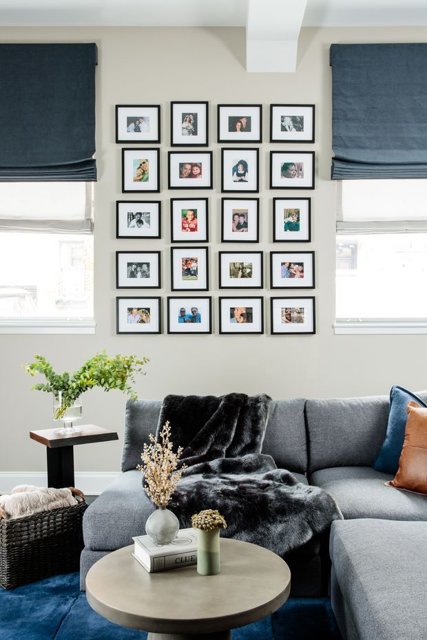 A bright, cosy living room with a large grey sectional sofa, black faux-fur throw, blue and tan accent cushions, and a round pale stone or concrete coffee table. A gallery wall of small black-framed family photographs is arranged between two windows with dark blue Roman blinds. The room includes a woven black basket, side table, vase of greenery, and layered neutral furnishings. A white ceiling beam and light neutral walls create a refined contemporary interior.