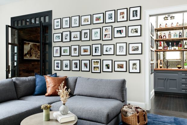 A refined living room with a large grey sectional sofa, blue and rust accent cushions, a gallery wall of framed artwork, and a dark-framed glazed doorway leading to a bar area with built-in cabinetry and open bottle shelving. Dark timber flooring and detailed dark joinery add contrast to the light walls.