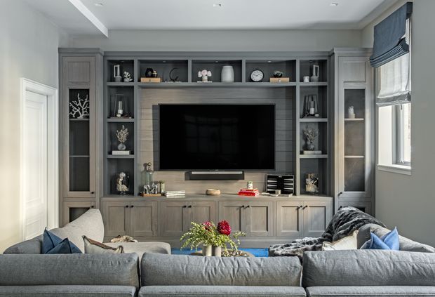 A refined living room centred on a large wall-mounted television and extensive built-in grey cabinetry. The joinery includes enclosed display towers, open shelving, lower cupboards, and a horizontal timber-toned feature backing. A large sectional sofa occupies the foreground, with blue cushions, soft furnishings, and a floral arrangement adding colour. The room has light grey walls, recessed ceiling lighting, white trim, and a Roman blind at the window.