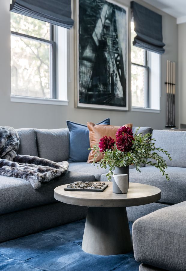 A refined living room with a large grey sectional sofa, blue and rust accent cushions, a round concrete coffee table, blue rug, and a floral arrangement. Tall windows with dark Roman blinds, a large framed artwork, and a modern decorative floor piece complete the room.