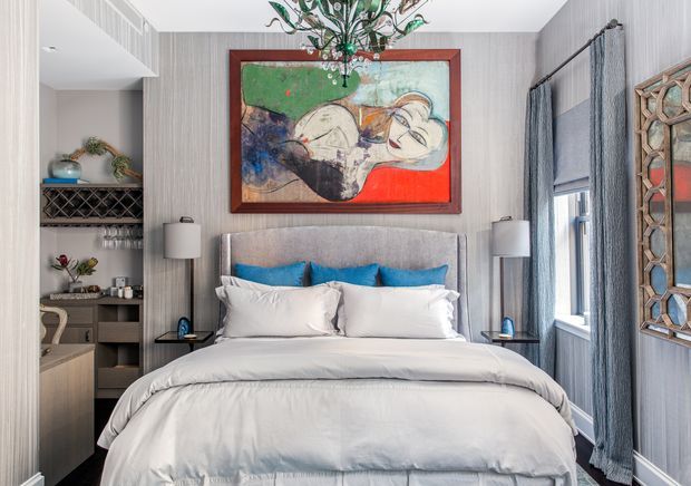 A refined bedroom with a large upholstered bed dressed in white bedding and accented by bright blue cushions. The room has vertically textured grey wallcovering, matching full-height curtains, symmetrical bedside tables and lamps, a decorative chandelier, a colourful framed artwork above the bed, and built-in shelving with glassware and decor. A mirrored or decorative geometric wall panel is visible on the right.