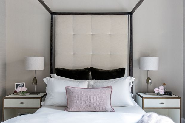 A refined bedroom centered on a large four-poster bed with a tall cream upholstered, tufted headboard. The bed is dressed in crisp white linens with dark accent cushions and a muted blush-pink lumbar pillow. Matching bedside tables with light drawers and dark trim sit beneath white table lamps, creating a symmetrical and polished composition.