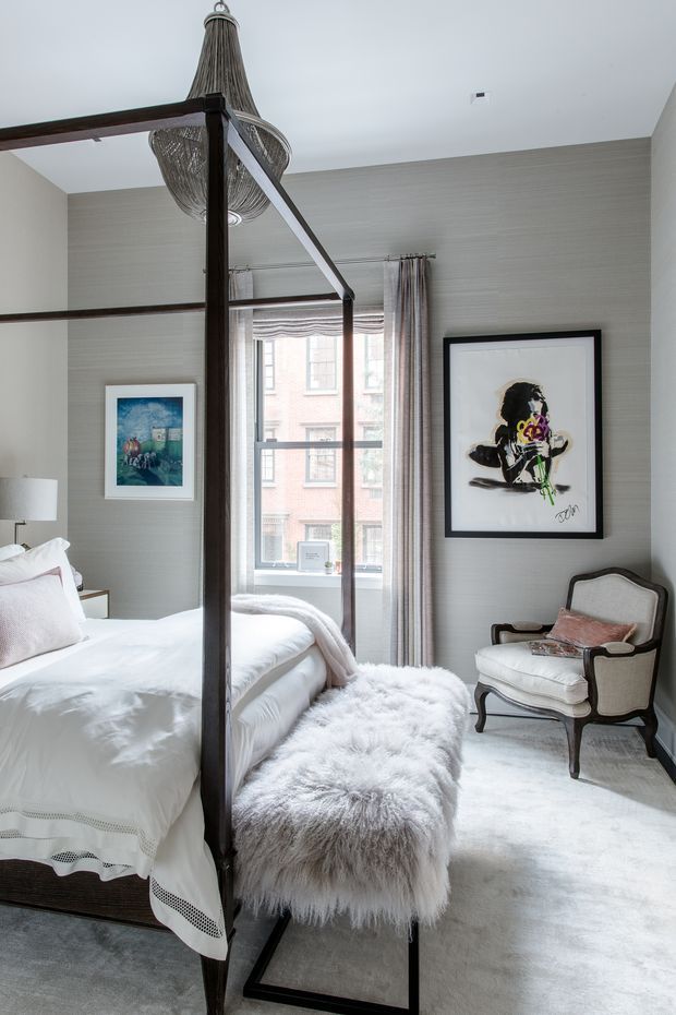 A bright, refined bedroom with a dark timber four-poster bed, white bedding, a grey upholstered bench topped with a fluffy throw, and a classic upholstered armchair. The room has textured pale grey walls, full-height curtains, a large window, framed artwork, and a sculptural woven pendant light. The palette is predominantly white, grey, and dark timber with soft pink accents.