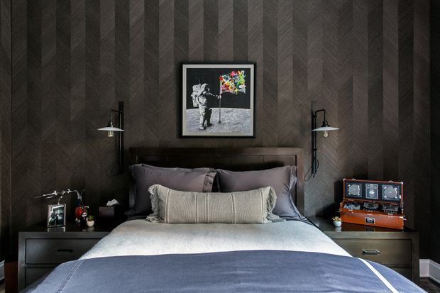 A dark, moody bedroom centered on a neatly made bed with layered grey and charcoal bedding, a padded dark headboard, and matching bedside tables. The walls feature dramatic dark herringbone wood panelling, with black wall-mounted lamps on either side of the bed. Framed artwork hangs above the headboard, and a vintage-style orange radio sits on the right bedside table.
