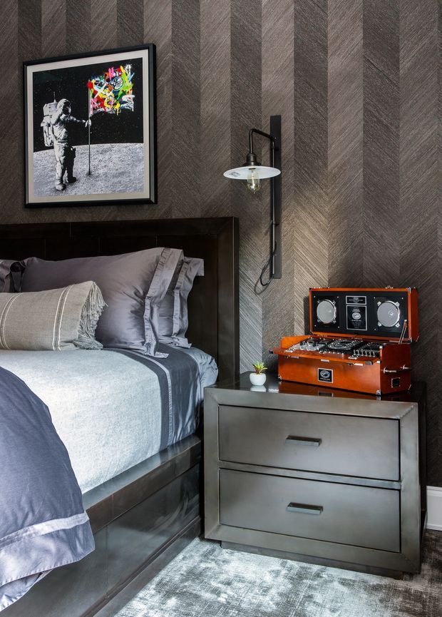 A moody, refined bedroom vignette featuring a dark upholstered or timber bed with layered grey bedding, a metallic two-drawer bedside table, a black wall-mounted swing-arm lamp, framed artwork, and a vintage orange-brown record player. The wall is finished in a dramatic dark herringbone-patterned surface, with a pale textured rug grounding the scene.
