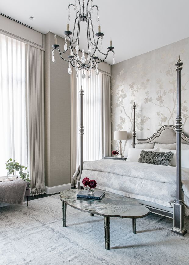 Elegant, bright bedroom with a tall ornate four-poster bed, pale upholstered bedding, patterned botanical feature wallpaper, floor-to-ceiling sheer curtains, a dramatic crystal chandelier, and a sculptural stone-topped coffee table over a light rug. The palette is predominantly soft white, grey, and beige with refined traditional detailing and subtle floral accents.