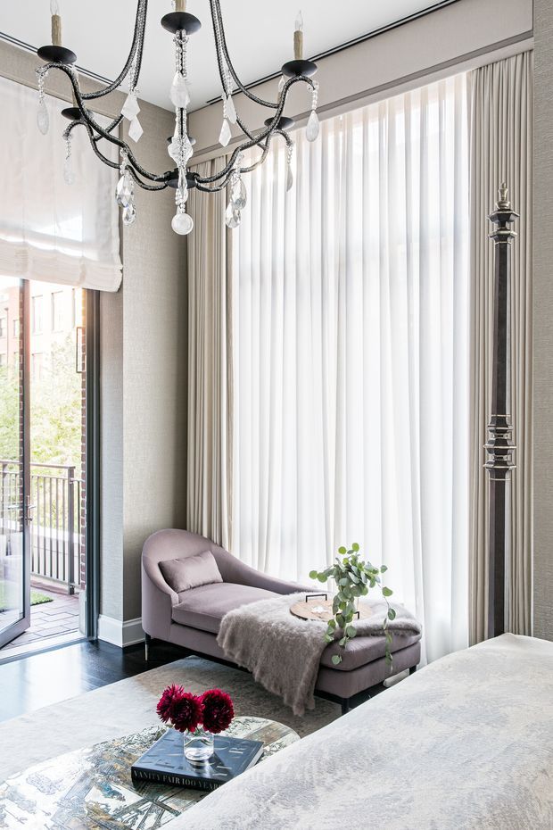 Elegant, light-filled sitting area with tall sheer curtains, taupe textured walls, dark timber flooring, and a large ornate crystal chandelier. A curved blush upholstered chaise sits beside full-height glazing opening to a balcony, while a pale patterned rug and low coffee table occupy the foreground. The space has a refined, softly glamorous aesthetic with high ceilings and generous natural light.