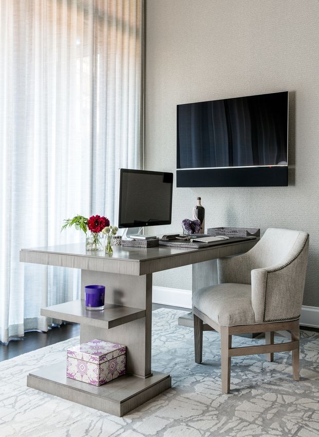 A refined home office with a contemporary desk positioned beside floor-to-ceiling sheer curtains. The desk has open shelving, a computer monitor, decorative accessories and flowers, while a textured upholstered chair sits in front. A wall-mounted television and soundbar are visible above the desk area. The room uses pale grey textured walls, light grey timber-look joinery, a patterned pale rug or carpet, and soft natural light.
