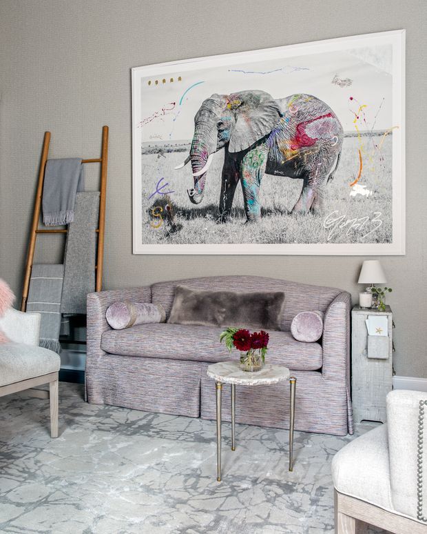 A refined living room with a soft grey upholstered sofa, pale armchairs, a small round marble-topped coffee table, patterned rug, and a leaning timber ladder with folded textiles. The room features textured grey walls, a white-framed oversized elephant artwork, metallic accents, and muted mauve, grey, and neutral tones. The styling is polished and eclectic with a playful art focal point.