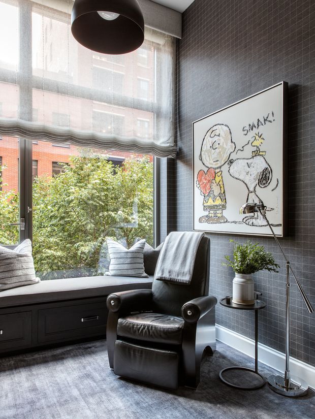 A compact, stylish sitting area with a black leather recliner beside a dark built-in window seat, layered with grey cushions and a throw. Floor-to-ceiling windows with sheer roller blinds overlook greenery and neighbouring brick buildings. The room features dark small-format tiled walls, grey carpeting, a large black pendant light, a slender floor lamp, and a framed Snoopy illustration with a potted plant on a small round side table.