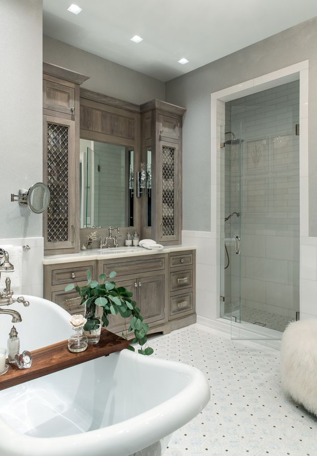 A spacious traditional-style bathroom with a freestanding white bathtub in the foreground, a large timber vanity with mirrored diamond-pattern cabinet doors, and a glass-enclosed walk-in shower. The room features grey walls, white subway tile shower walls, white trim, and patterned light tile flooring. Recessed ceiling lights and polished metal fixtures complete the refined, classic look.