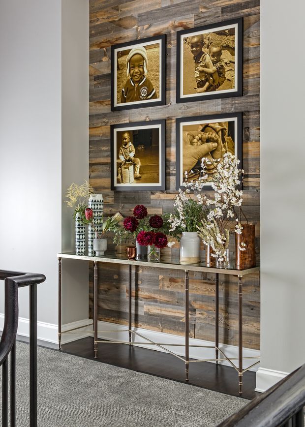 A refined hallway or entryway vignette featuring a narrow metal-framed console table against a feature wall clad in reclaimed timber planks. Four sepia-toned framed photographs hang above the console, which is styled with ceramic vessels, flowers, and decorative objects. Dark timber flooring, a grey area rug, white skirting boards, and neutral painted walls complete the space.