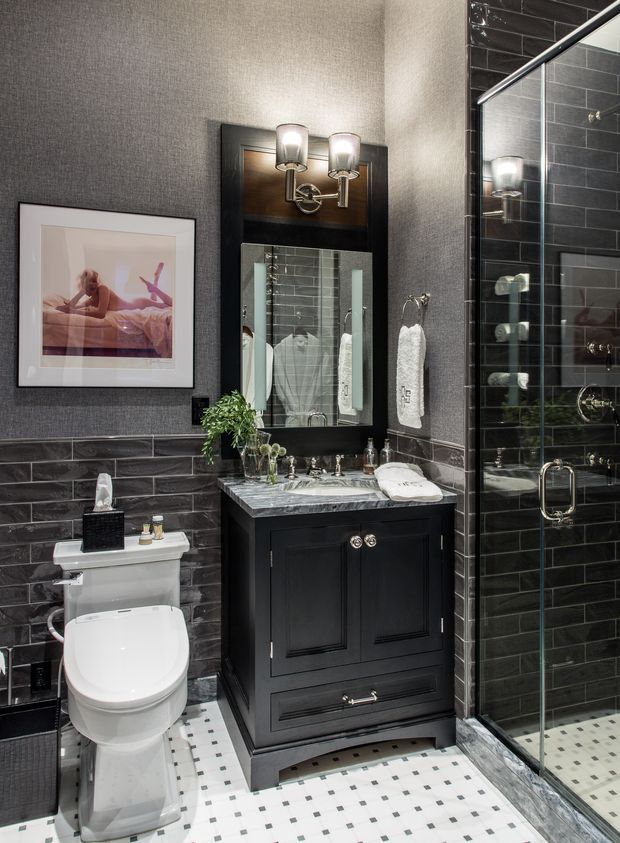A compact, richly detailed bathroom with dark charcoal subway-tile walls, a white mosaic tile floor, black traditional vanity cabinetry, a marble countertop, and a framed mirror with twin wall sconces. A toilet, pedestal-style basin vanity, towels, and a glass-enclosed shower are clearly visible. The space has a refined, high-contrast, classic look with metallic accents and framed artwork.