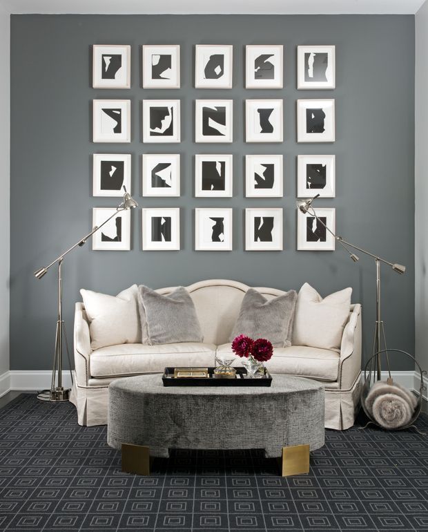 A refined living room with a curved cream sofa against a charcoal-grey feature wall, arranged with grey and white cushions, a large rounded upholstered ottoman, and contemporary slim floor lamps. A grid of framed black-and-white abstract artworks decorates the wall, while a dark geometric patterned carpet and small floral arrangement add contrast. The room has a polished, tailored and slightly glamorous feel.