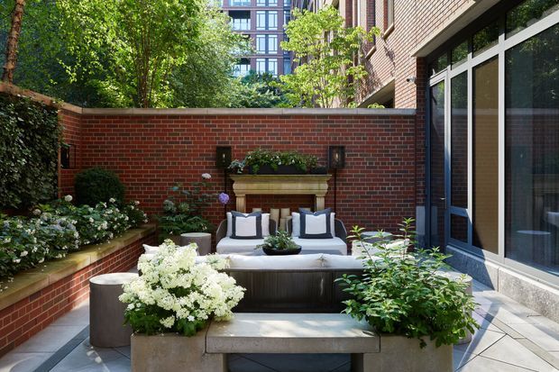 A refined urban courtyard terrace enclosed by red brick walls and a brick-clad building facade. The space features built-in raised garden beds, abundant greenery and white flowering plants, two lounge chairs, a dark outdoor coffee table, and a stone bench. Large dark-framed glazed doors or windows open onto the courtyard, while a taller apartment building is visible beyond the wall.