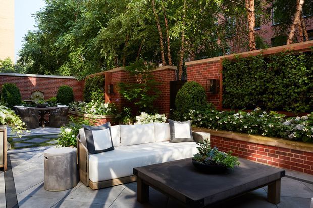 A landscaped urban courtyard terrace with a contemporary outdoor seating area. A pale upholstered sofa, dark low coffee table, and metal side table sit on large grey paving slabs, surrounded by lush planted borders, clipped shrubs, white flowering plants, and mature trees. Red brick garden walls, raised brick planters, and dense greenery define the enclosed outdoor space.