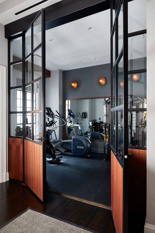 A home gym viewed through open black-framed glass-and-timber doors. The room contains cardio equipment, a multi-station weight machine, dark rubber flooring, a large wall mirror, and warm circular wall lights. Rich dark wood flooring and reddish timber door panels contrast with charcoal grey walls and white ceilings, creating a refined, moody gym interior.
