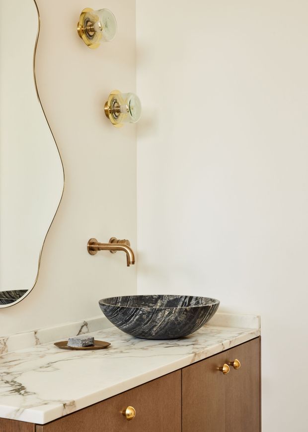 Close-up of a refined bathroom vanity with a dark veined stone vessel basin set on a pale marble countertop. The warm timber cabinetry has small brass-gold knobs, complemented by a brass wall-mounted faucet and matching glass wall sconces. An organic curved-edge mirror is partially visible on the left, while the light neutral walls create a calm, luxurious atmosphere.