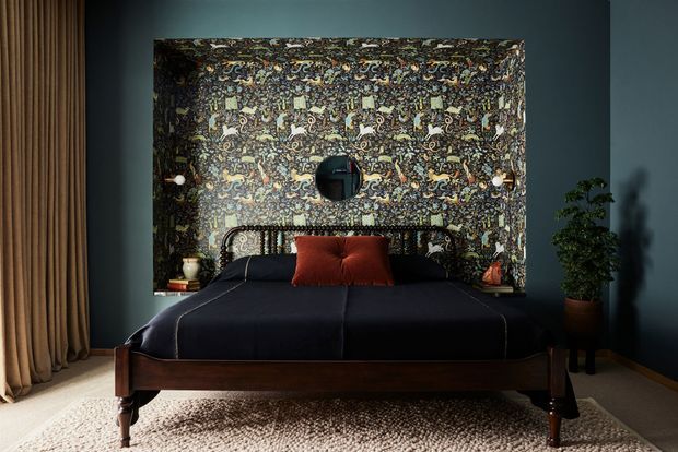 A moody, richly styled bedroom viewed directly from the foot of the bed. The room features a dark upholstered bed with a warm dark-wood timber frame and tapered legs, a patterned decorative feature wall behind the headboard, dark blue-grey painted walls, full-height beige curtains, a rust-red accent cushion, a round wall mirror, and a potted plant. The overall composition is symmetrical and dramatic, with eclectic patterned detailing and warm timber accents.