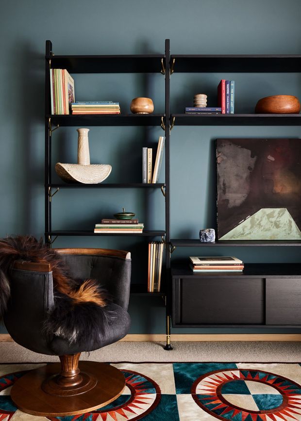 A richly styled living room featuring a dark teal painted wall with black open shelving and integrated low cabinetry. The shelving displays books, ceramics, sculptures and decorative objects, while a black leather lounge chair with a fur throw sits in the foreground. A large abstract artwork, patterned geometric rug, timber chair base and warm wood accents add contrast. The overall look is moody, refined and eclectic with mid-century influences.