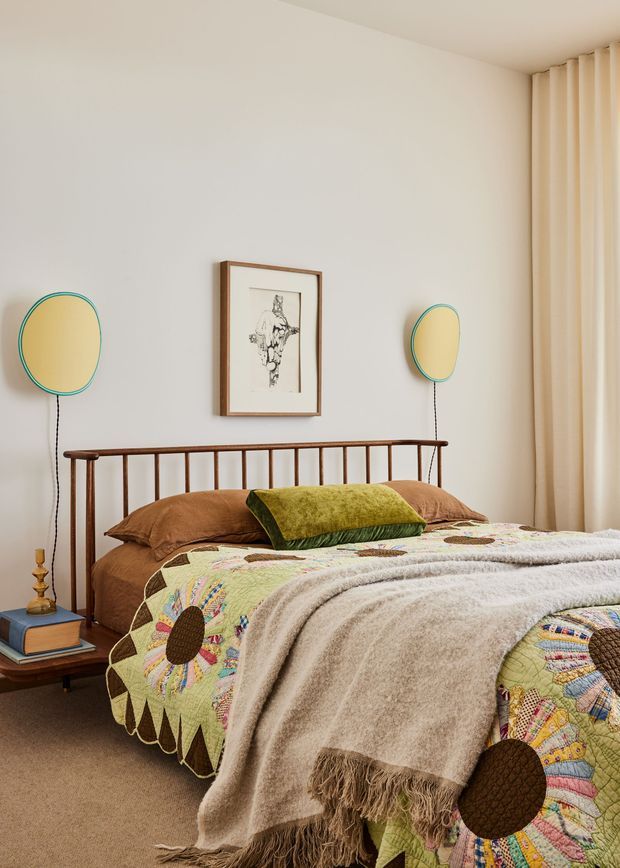 A warm, softly lit bedroom featuring a wooden spindle bed with layered brown bedding, a green accent cushion, a colourful patchwork quilt, and a textured beige throw. Cream curtains cover the right side of the room, while retro oval wall lights and framed artwork decorate the pale wall. A small wooden bedside table with books and a lamp sits beside the bed.