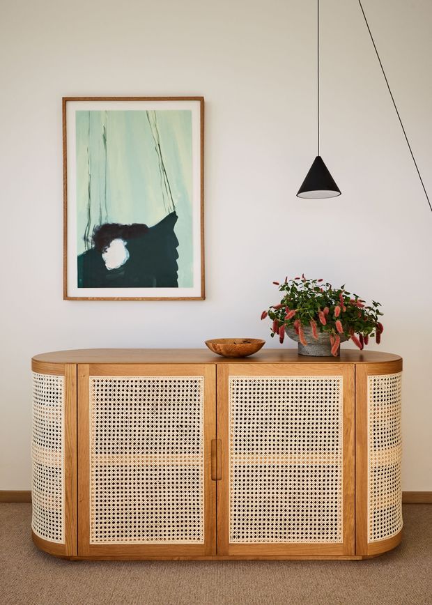 A warmly styled interior vignette featuring a mid-century-inspired timber sideboard with rounded ends and woven cane door panels. The sideboard stands against a soft neutral wall above a brown carpet, accompanied by a framed abstract artwork, a black pendant light, a wooden bowl, and a leafy potted plant.