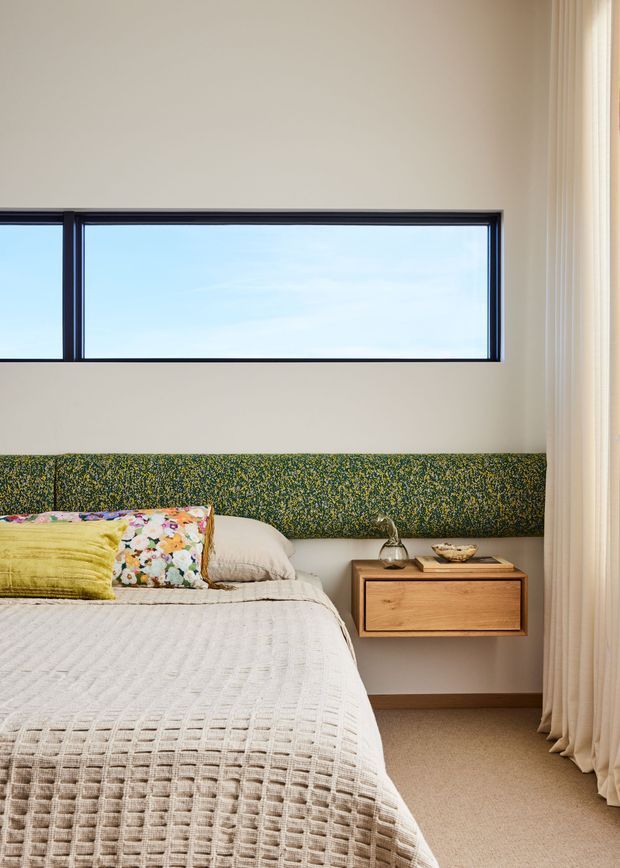 A bright, softly styled bedroom with a low platform bed, textured cream bedding, layered floral and yellow cushions, and a long upholstered green patterned headboard. A floating timber bedside cabinet with a small lamp and bowl sits beside the bed. A wide horizontal black-framed window provides natural light, while full-height cream curtains frame the right side. The room has a calm, warm, contemporary character with restrained mid-century-inspired details.