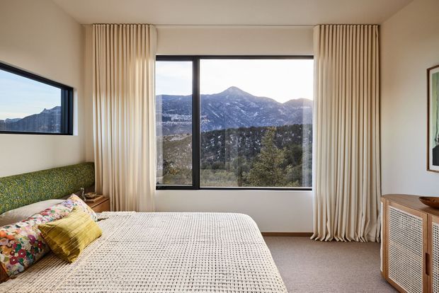 A serene bedroom with a large central picture window overlooking a rugged mountain landscape. The room features floor-to-ceiling cream curtains, a textured upholstered green headboard, colourful patterned cushions, a mustard accent cushion, a white textured bedspread, neutral walls and carpet, and a woven-front timber bedside cabinet. A smaller side window and framed artwork are also visible.