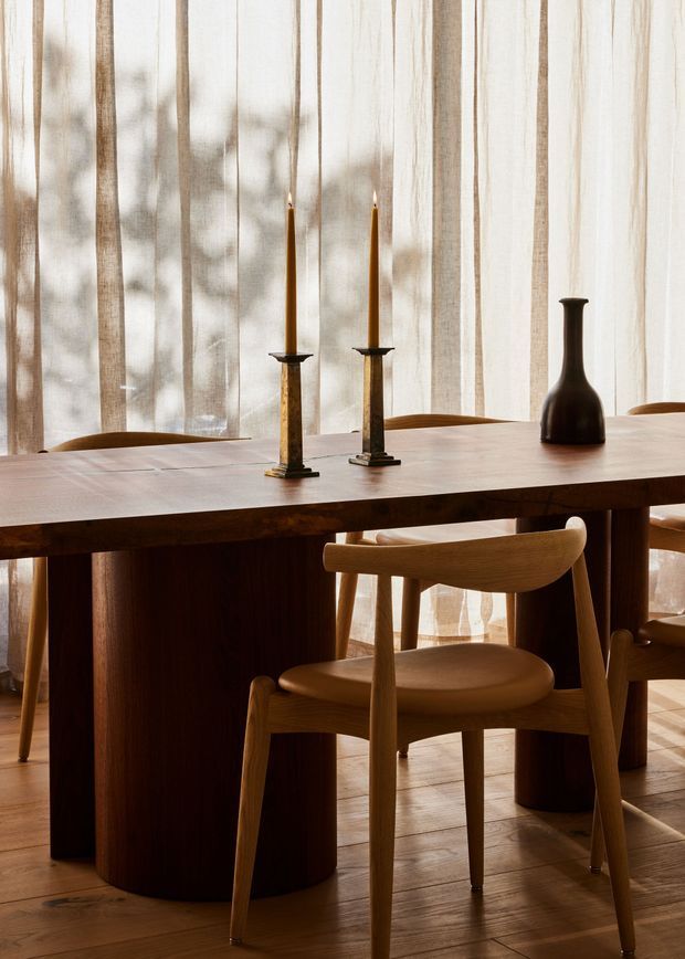 A warmly lit dining area centered on a substantial dark timber dining table with rounded pedestal supports and matching wooden chairs. Tall sheer curtains diffuse daylight across the room, while two slender candles in dark holders and a dark sculptural vase form a restrained tabletop arrangement. Timber flooring and predominantly natural wood tones create an intimate, refined atmosphere.
