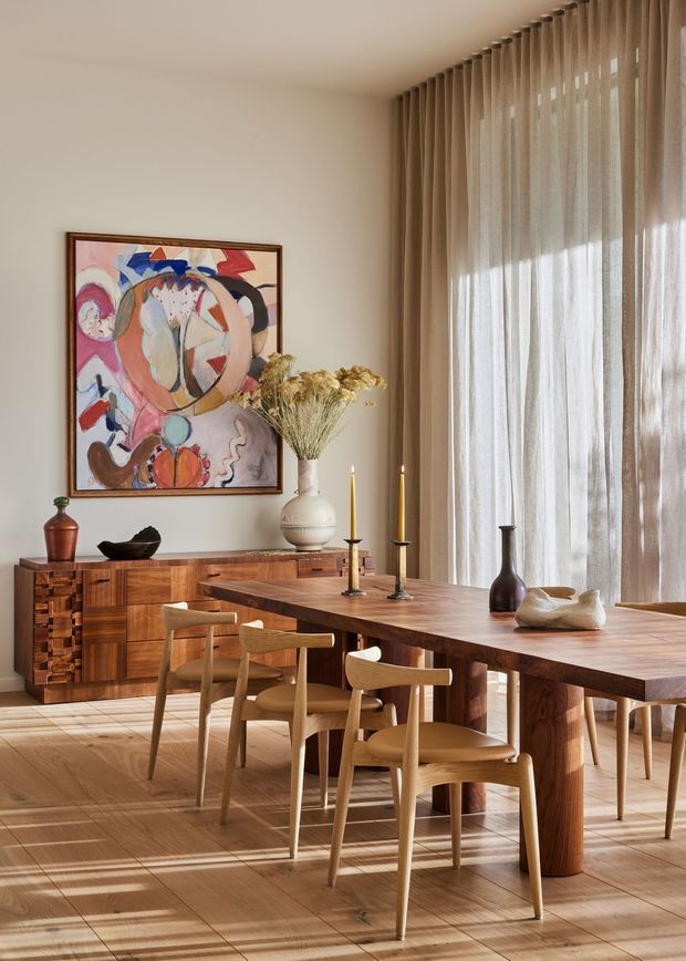A warm, refined dining room with a long solid timber dining table and matching sculptural timber chairs. Floor-to-ceiling sheer curtains line the right side, allowing abundant natural light into the room. A richly patterned timber sideboard sits against the wall beneath a colourful abstract artwork, with a ceramic vase of dried flowers and understated candle and ceramic decorations on the table. The room has timber flooring, neutral plastered walls, and a calm contemporary natural-materials aesthetic.