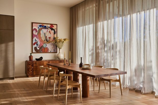 Warm, refined dining room with a large solid-wood dining table and matching timber chairs, set against full-height sheer curtains and expansive glazing. A timber sideboard, abstract artwork, sculptural ceramic vessels, and soft natural light create an inviting contemporary interior. Large-format light beige floor tiles and neutral walls complete the space.