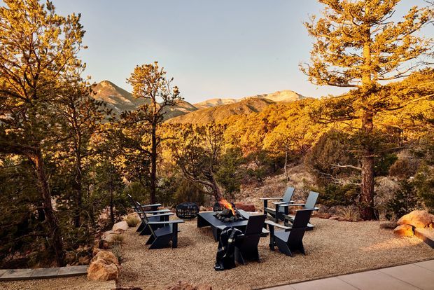 An outdoor terrace or patio set within a forested mountain landscape. A gravel-paved seating area contains dark modern lounge chairs arranged around a central fire pit, with additional seating and a low stone edge. Autumn-colored trees frame views of rugged mountain peaks in the background.