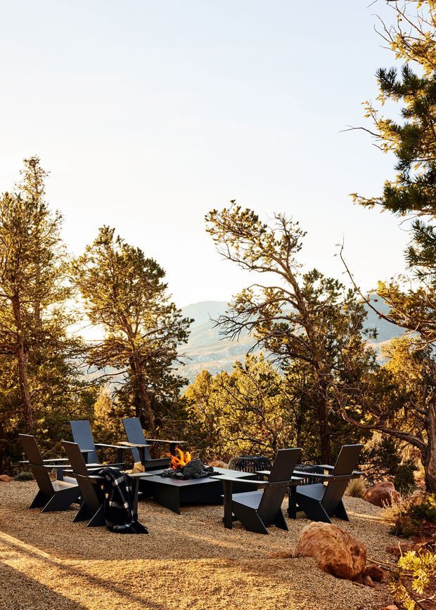 Outdoor lounge and dining area set on a gravel terrace among mature trees, with angular dark outdoor chairs arranged around a low rectangular fire table and a visible open flame. The setting overlooks a distant mountainous landscape and is lit by warm, low-angle sunlight.