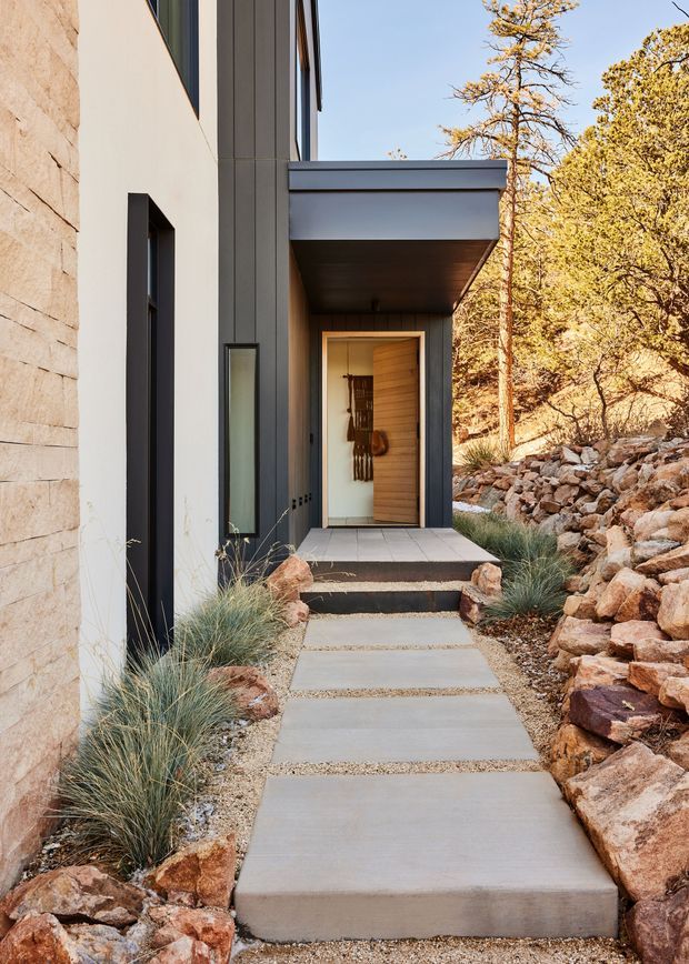 A modern residential entry viewed from an exterior walkway. The facade combines pale rendered walls, dark vertical cladding, and a light natural-stone wall. A projecting flat-roof canopy shelters a timber-lined front door, with a narrow vertical window beside it. Concrete stepping slabs set in gravel lead through a dry, rocky landscaped garden with ornamental grasses and mature trees.