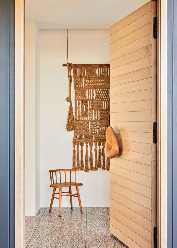 A warmly lit entryway viewed through an open horizontal timber-planked door. The compact vestibule has white walls, terrazzo-style flooring, a vintage wooden spindle-back chair, and a large handwoven macramé wall hanging in natural brown fibres. The door and surrounding frame are natural timber, with dark hinges and a sculptural wooden handle. Blue-grey exterior cladding is visible at the sides.