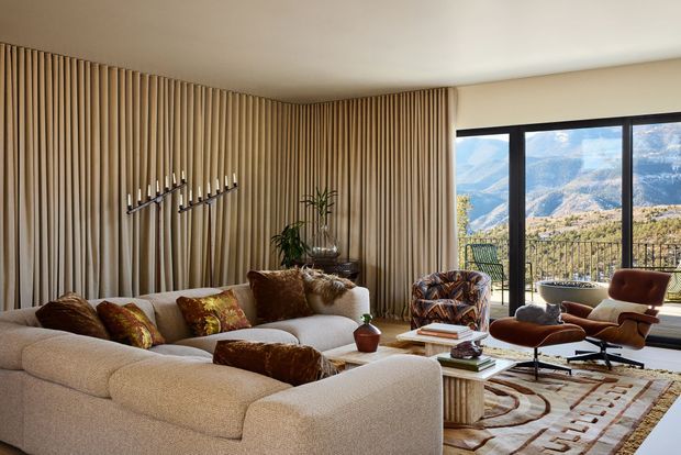 A warm, refined living room with a large curved cream sectional sofa, sculptural lounge chairs, patterned cushions, a low coffee table, and a geometric area rug. The room features a full-height vertical timber slat feature wall, neutral plaster ceiling, dark-framed floor-to-ceiling glazing, and expansive mountain views beyond the balcony. A minimalist linear wall light and indoor plant add visual interest.
