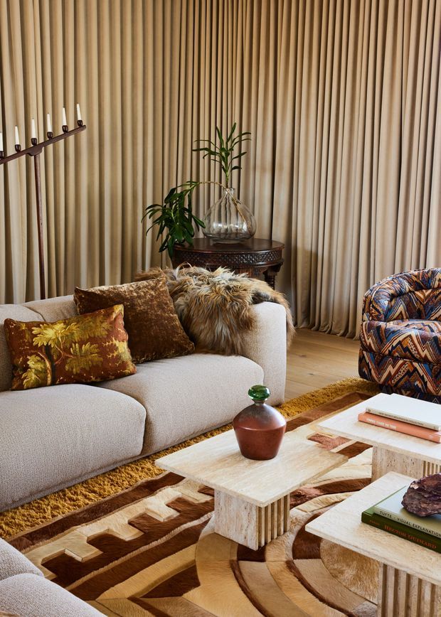 A richly layered living room vignette with a curved light beige sectional sofa, patterned and textured cushions, a bold geometric brown-and-cream rug, sculptural pale stone coffee tables, and a vividly patterned accent armchair. Full-height pleated beige curtains cover the wall, while a dark round side table, glass vase with greenery, candles, and warm timber flooring add depth. The overall look is warm, eclectic, and refined with strong textural and geometric detailing.