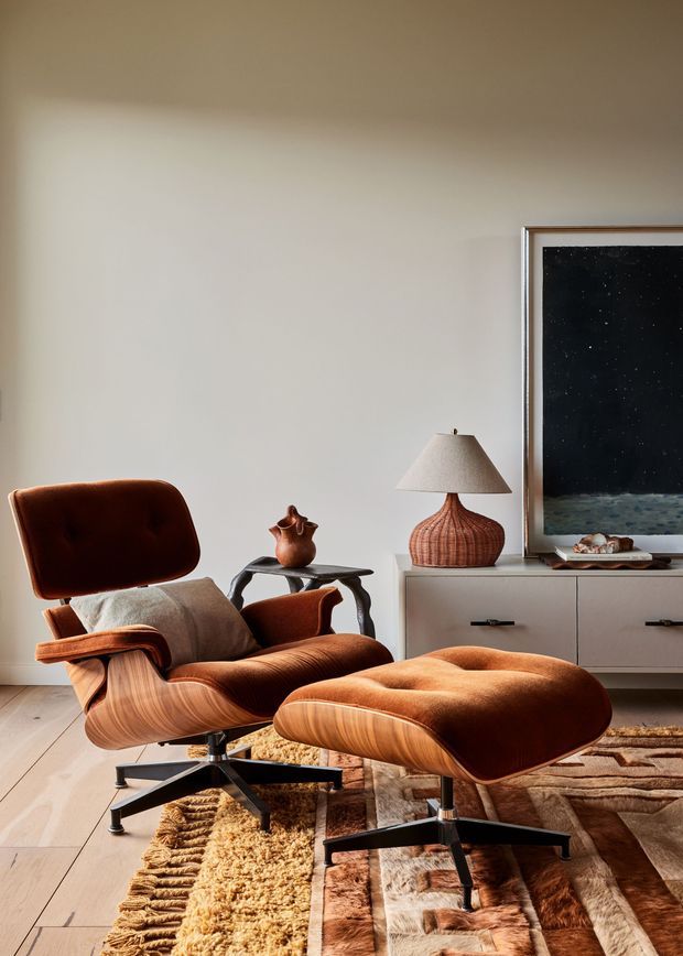 A warmly styled mid-century-inspired living room featuring a sculptural brown leather lounge chair with matching ottoman, a low timber side table, a textured table lamp, and a minimalist low-profile cabinet. A large framed artwork leans against the wall, while layered natural-toned rugs and timber flooring create a rich, inviting atmosphere.