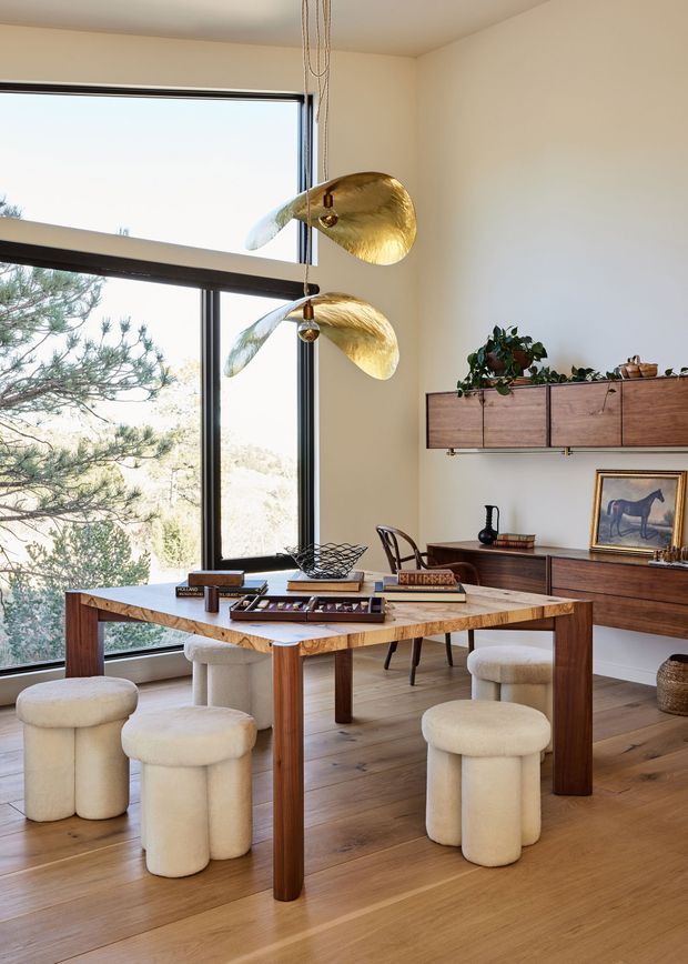 A warm, refined dining room with a large timber dining table surrounded by upholstered cream stools and chairs. Floor-to-ceiling corner glazing provides views of trees and a bright natural setting. The room includes mid-century-inspired timber wall cabinetry and a sideboard, sculptural gold pendant lights, indoor plants, and curated decorative objects. The palette is dominated by warm timber, cream, beige, and gold tones.