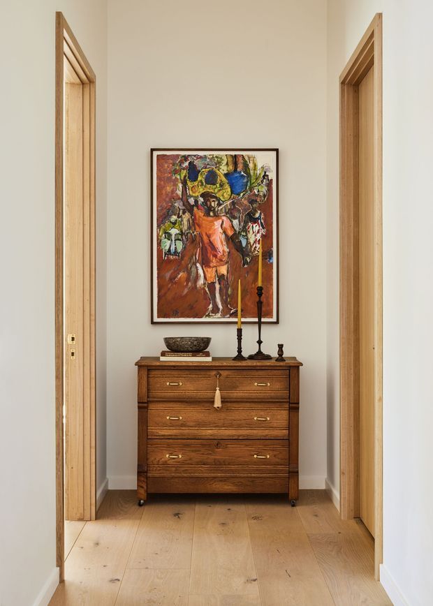 A narrow, softly lit hallway or entry alcove with pale timber floorboards, white walls, and natural timber door frames on both sides. A dark mid-century-style wooden chest of drawers is centered against the back wall, topped with decorative objects and candlesticks, with a colourful framed artwork above it. The space has a warm, refined, uncluttered character.