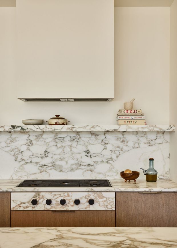 A close-up view of a refined kitchen cooktop and cabinetry wall. The kitchen features dramatic veined white marble across the splashback, benchtop, and drawer fronts, paired with warm dark timber cabinetry. A concealed rangehood, integrated black cooktop, brass-toned hardware, and a few decorative cooking and pantry objects complete the understated composition.
