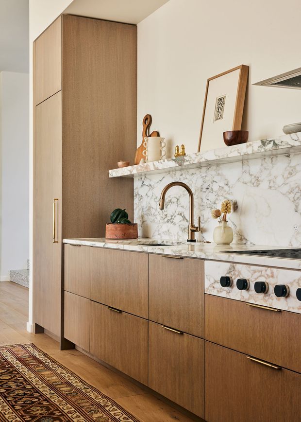 A warm, refined kitchen with full-height and base cabinetry in medium-toned timber, white veined marble-style benchtops and splashback, and brushed brass hardware. A bronze-toned gooseneck tap, integrated cooktop and oven, open shelf styling, framed artwork, and decorative objects add character. Timber flooring and a patterned runner reinforce the welcoming, sophisticated interior.