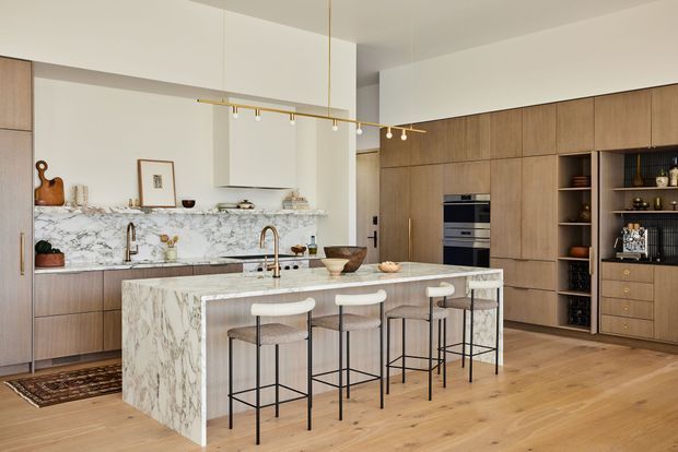 A spacious, refined contemporary kitchen with a large marble-clad island and integrated seating for four. Warm timber cabinetry spans the rear wall and incorporates a concealed appliance bank, open shelving and a dark gridded display niche. The kitchen features white upper cabinetry, a marble-look splashback, brass-toned tapware and linear pendant lighting, with light timber flooring and a neutral, softly warm palette.