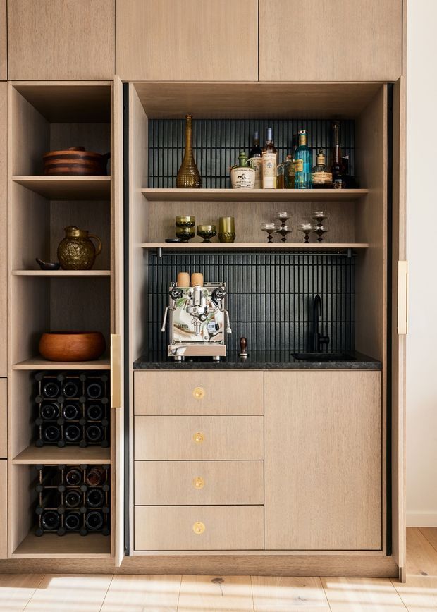 A built-in timber drinks station with open shelving, bottle storage, a dark ribbed backsplash, stone benchtop, sink and tap, espresso machine, glassware, and liquor bottles. The cabinetry is warm light wood with minimalist slab fronts and brass-toned hardware, creating a refined home bar or beverage nook.