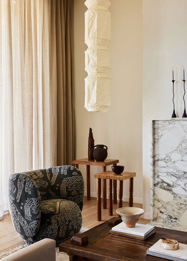 A warm, refined living room vignette with full-height sheer and taupe curtains, a sculptural upholstered lounge chair in a dark patterned fabric, nested wooden side tables, a rustic dark timber coffee table, ceramic vessels, and a large white marble fireplace surround. A dramatic hanging sculptural light adds visual interest against the cream walls.