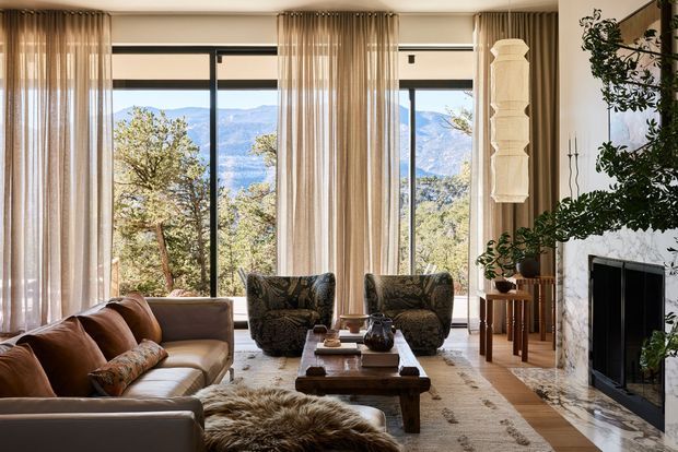 Elegant living room with a neutral upholstered sofa, patterned armchairs, a rustic timber coffee table, and a large textured rug. Full-height windows with sheer curtains open to a wooded mountain landscape. The room includes a sculptural pendant light, indoor greenery, and a substantial fireplace with a pale stone surround, creating a warm, refined, nature-connected interior.