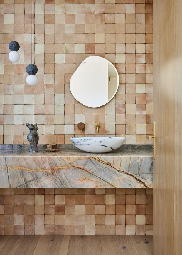 A warm, highly detailed bathroom vanity with a floating marble washbasin set on a dramatic veined stone counter. The wall is clad in small handmade-looking square tiles in varied terracotta, blush, cream and beige tones. A round mirror, spherical pendant lights, brass tapware and a sculptural faucet or decorative object add refined character. Timber cabinetry or wall panelling frames the right side, with timber flooring below. The composition has a boutique, earthy and luxurious feel.